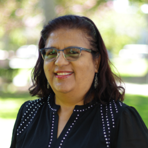 Woman smiling outdoors wearing blue-framed glasses and a dark dotted blouse; sunlight and greenery in the blurred background.